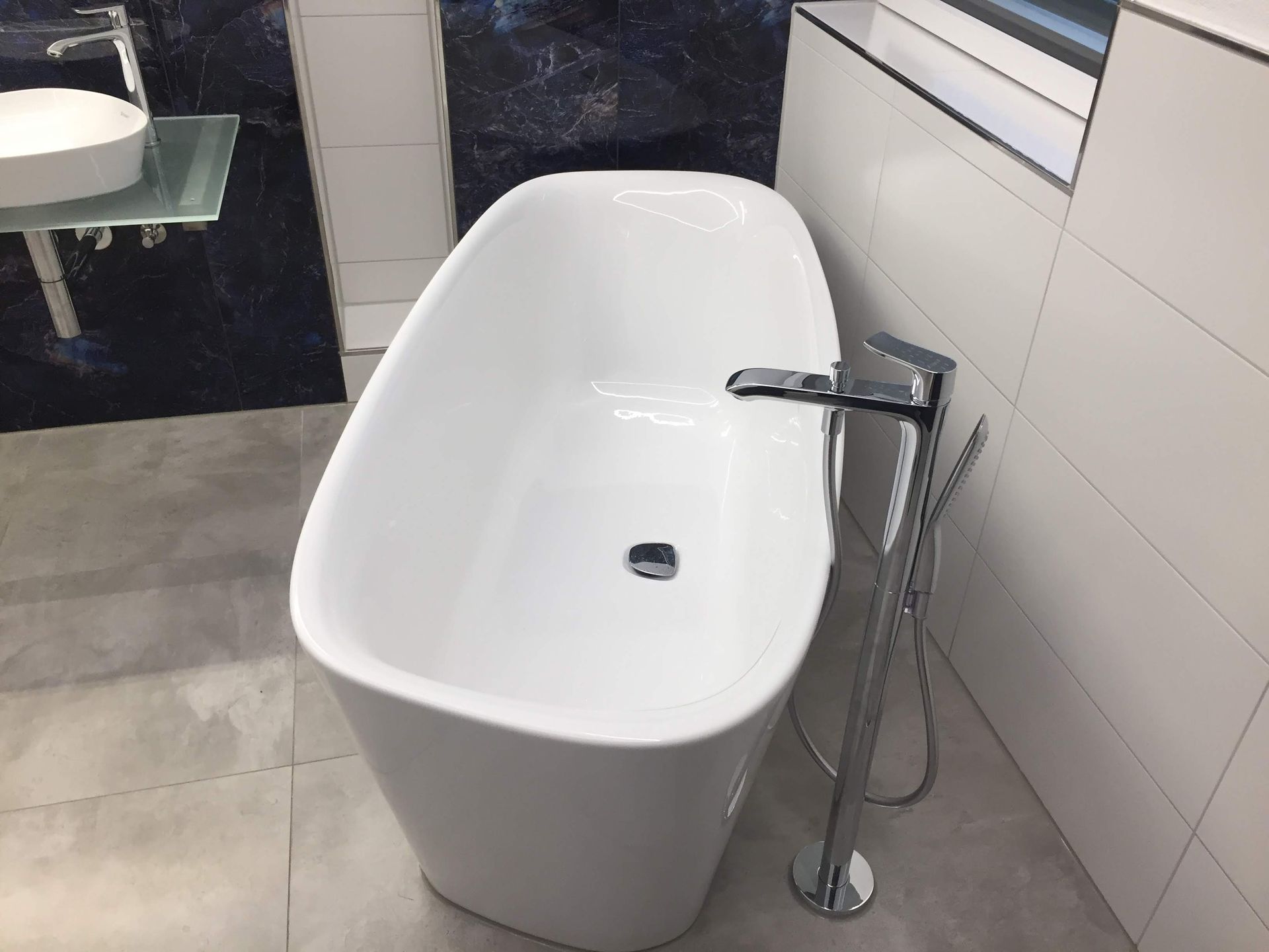 Freistehende weiße Badewanne mit bodenstehender Chromarmatur vor dunkler Fliesenwand